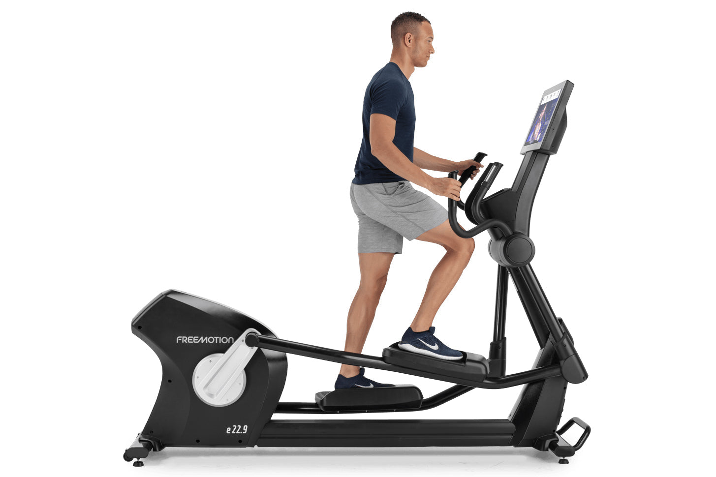 Man wearing navy t-shirt and gray shorts using black Freemotion e22.9 elliptical machine with digital display on white backgr