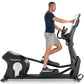Man using black Freemotion elliptical machine with digital display, wearing navy t-shirt and gray shorts on white background