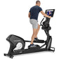 Man wearing navy t-shirt and gray shorts using black Freemotion e22.9 elliptical machine with tablet display showing video co