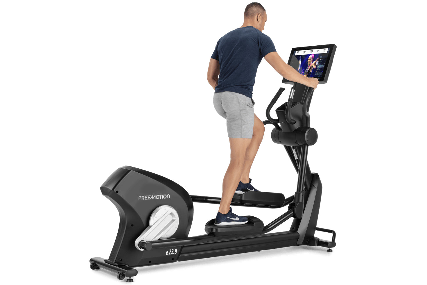 Man wearing navy t-shirt and gray shorts using black Freemotion e22.9 elliptical machine with tablet display showing video co