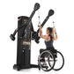 Black dual cable crossover machine with overhead pulleys, used by woman in wheelchair wearing navy tank top on white backgrou