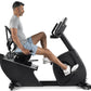 Man in gray shirt and blue shorts using black recumbent exercise bike with digital display on white background