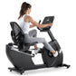 Woman in white tank top and gray leggings using black recumbent exercise bike with digital touchscreen display on white backg