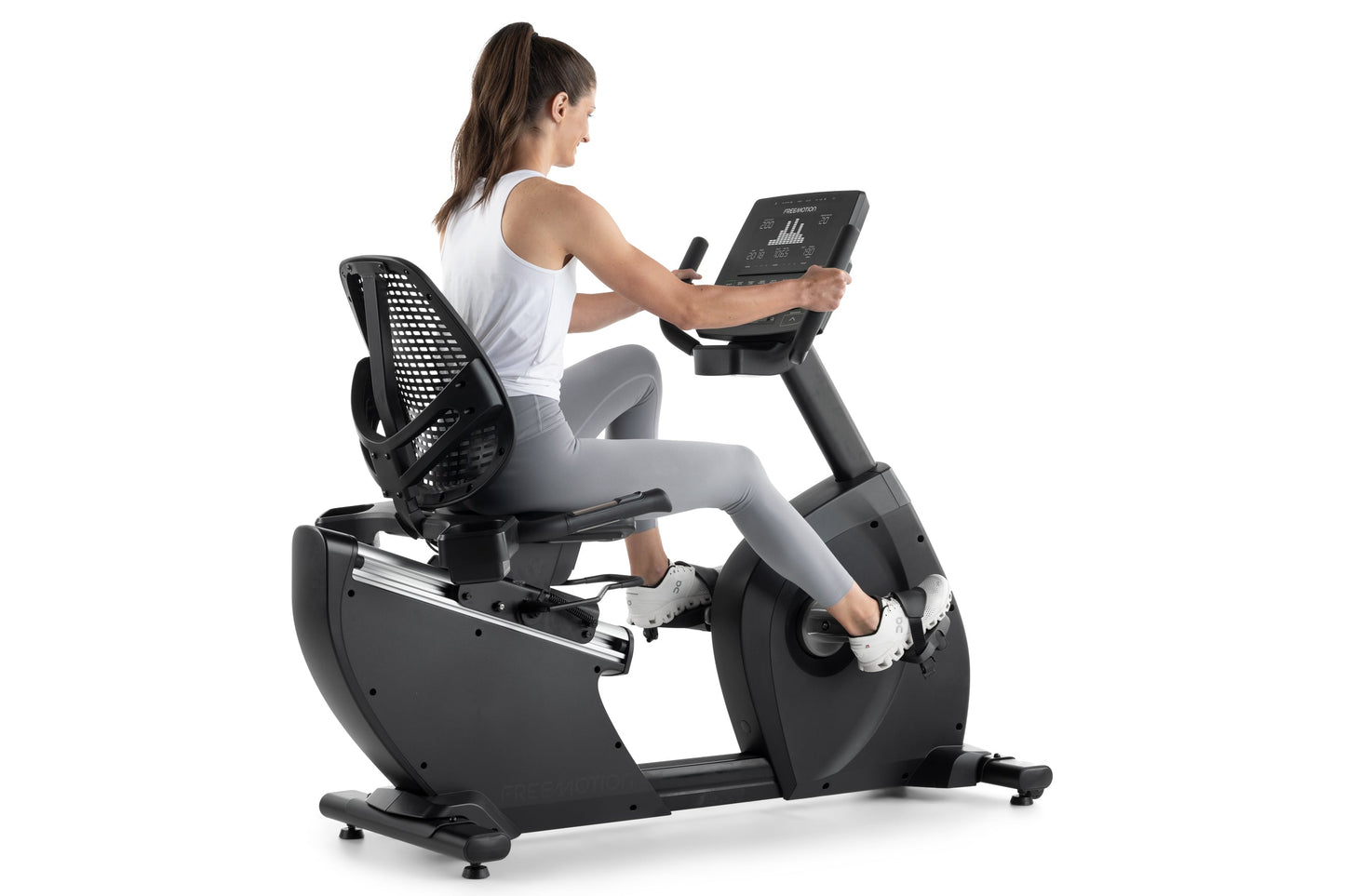 Woman in white tank top and gray leggings using black recumbent exercise bike with digital touchscreen display on white backg