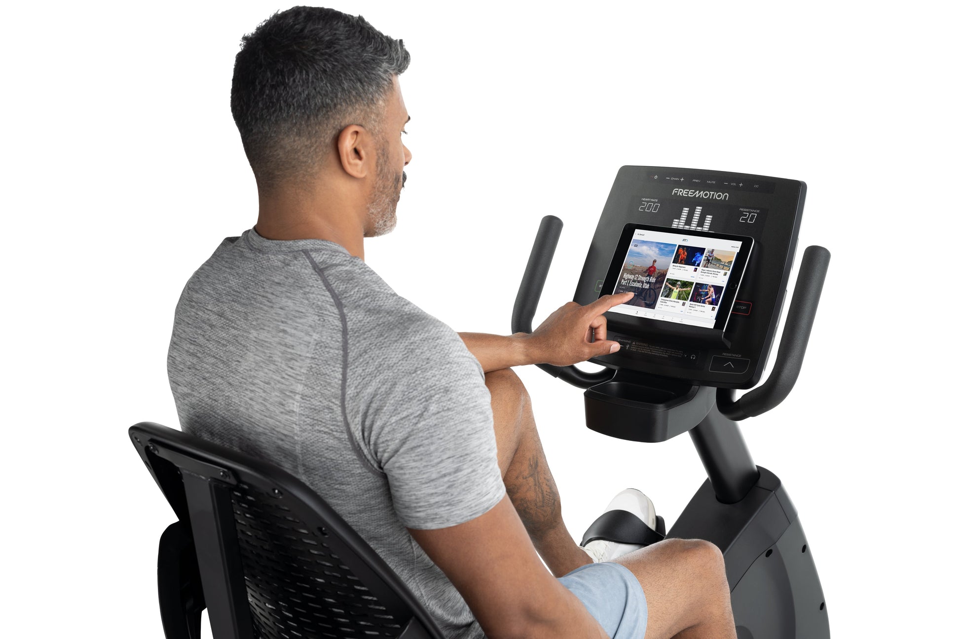 Man using black Freemotion recumbent exercise bike with touchscreen display showing workout content, worn by model on white b