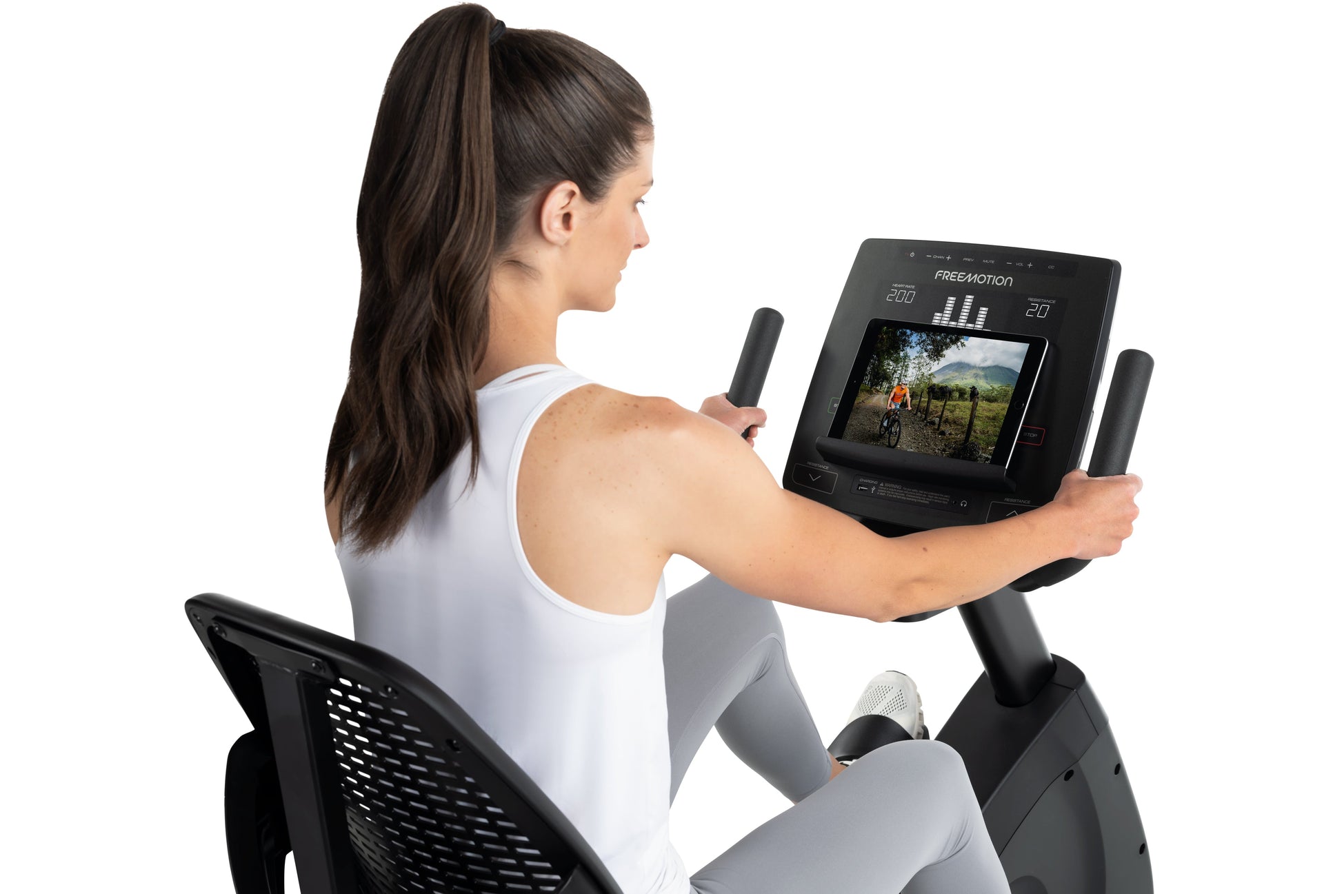 Woman using FreeMotion recumbent exercise bike with digital display showing outdoor biking video, worn white tank top and gra