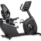 Black recumbent exercise bike with mesh backrest, digital console display, and pedal straps on white background