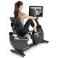 Woman using black Freemotion recumbent exercise bike with tablet display showing workout video, on white background
