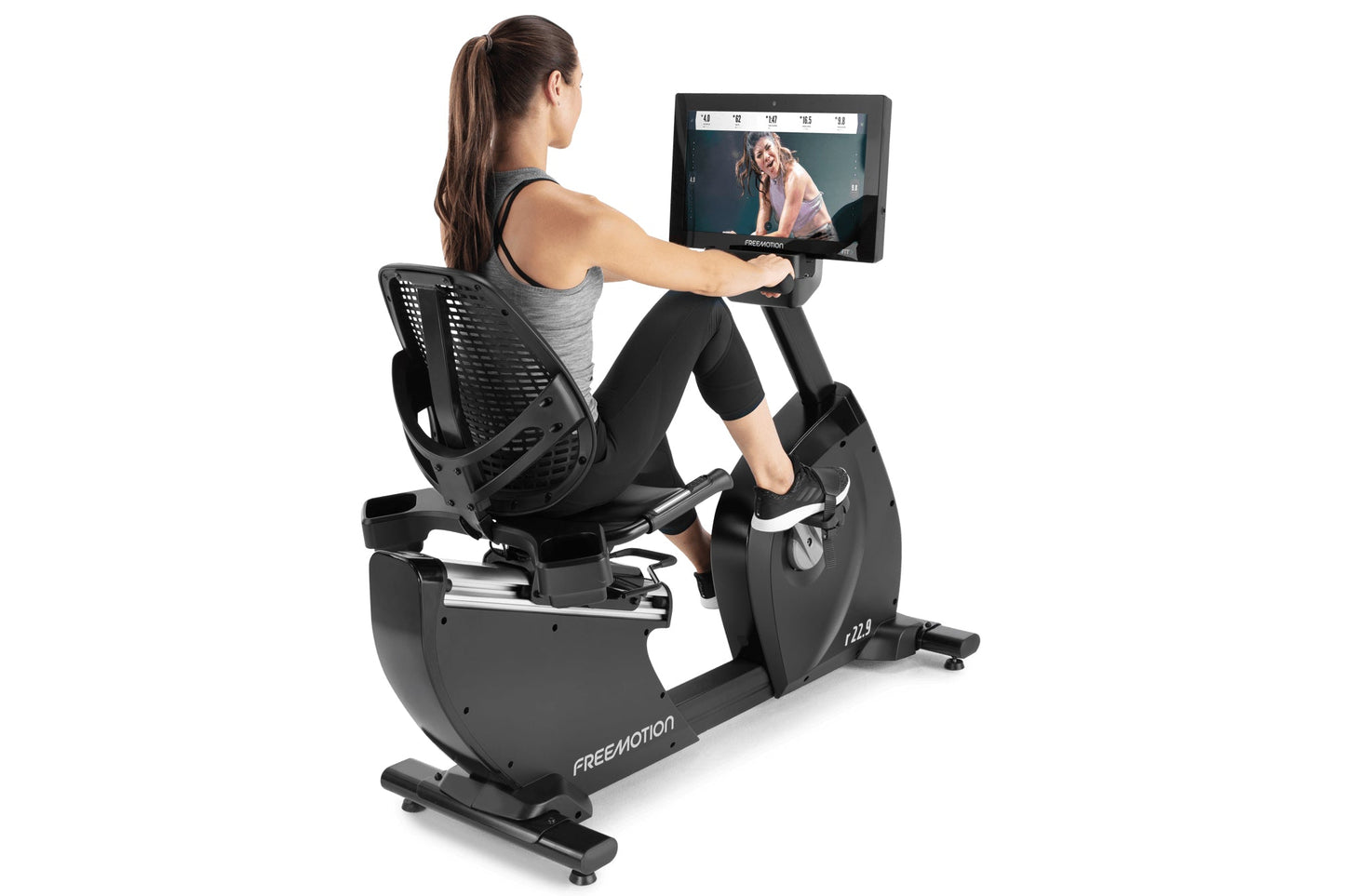 Woman using black Freemotion recumbent exercise bike with tablet display showing workout video, on white background