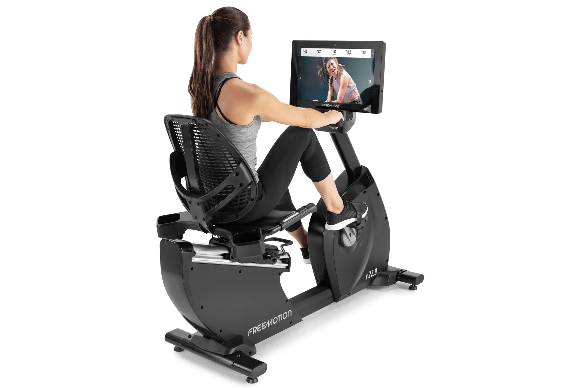 Woman using black Freemotion recumbent exercise bike with tablet display showing workout video, on white background