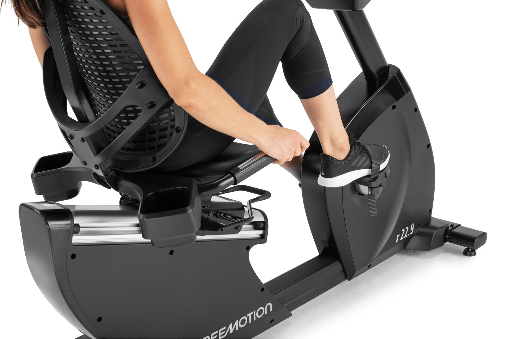 Person using black Freemotion r22.9 recumbent exercise bike with mesh backrest and foot pedals on white background