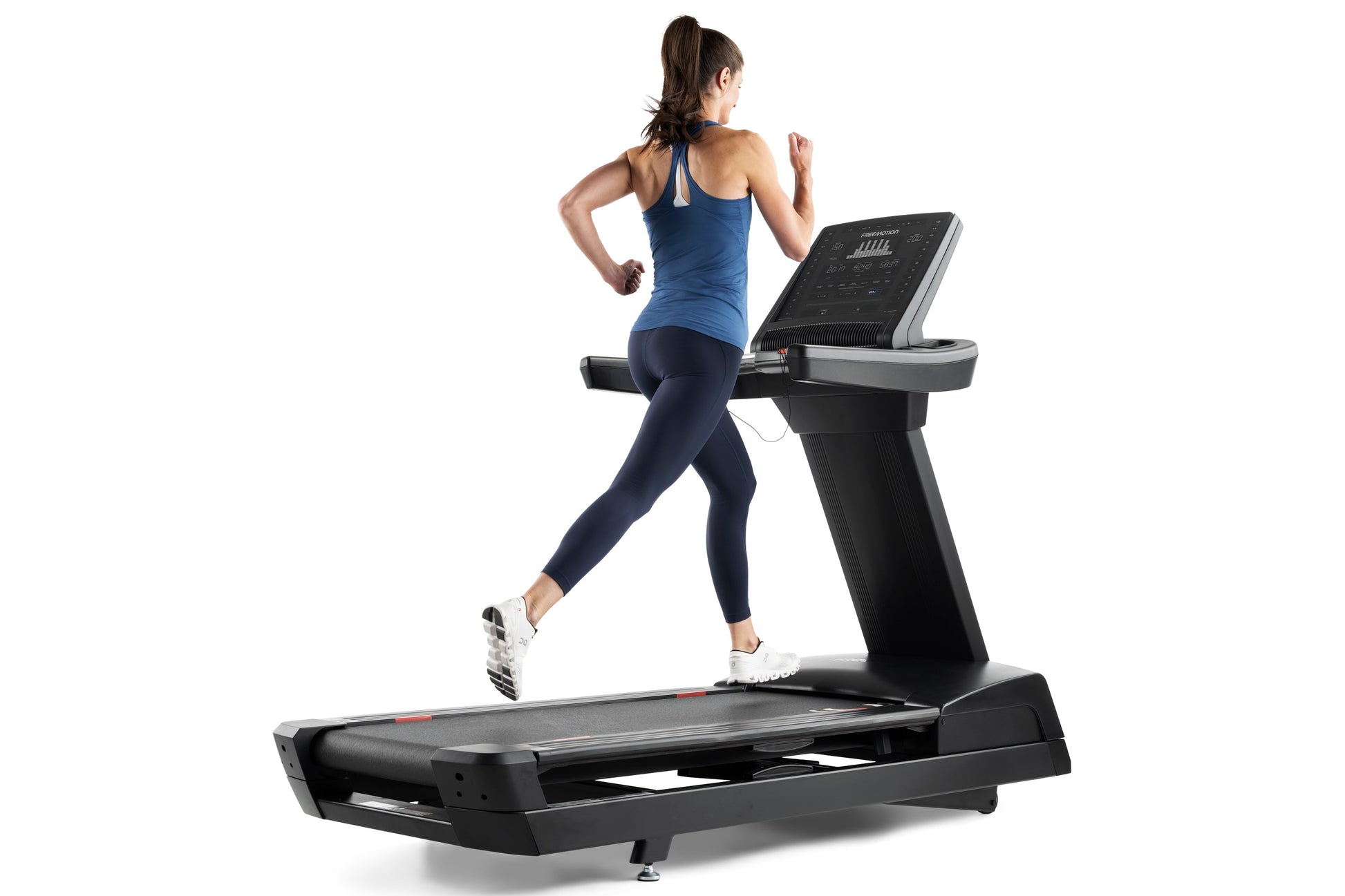 Woman running on black Freemotion treadmill wearing blue halter tank top and navy leggings, viewed from side on white backgro