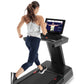Woman in gray tank top and navy capris running on black Freemotion treadmill with touchscreen display showing workout class i