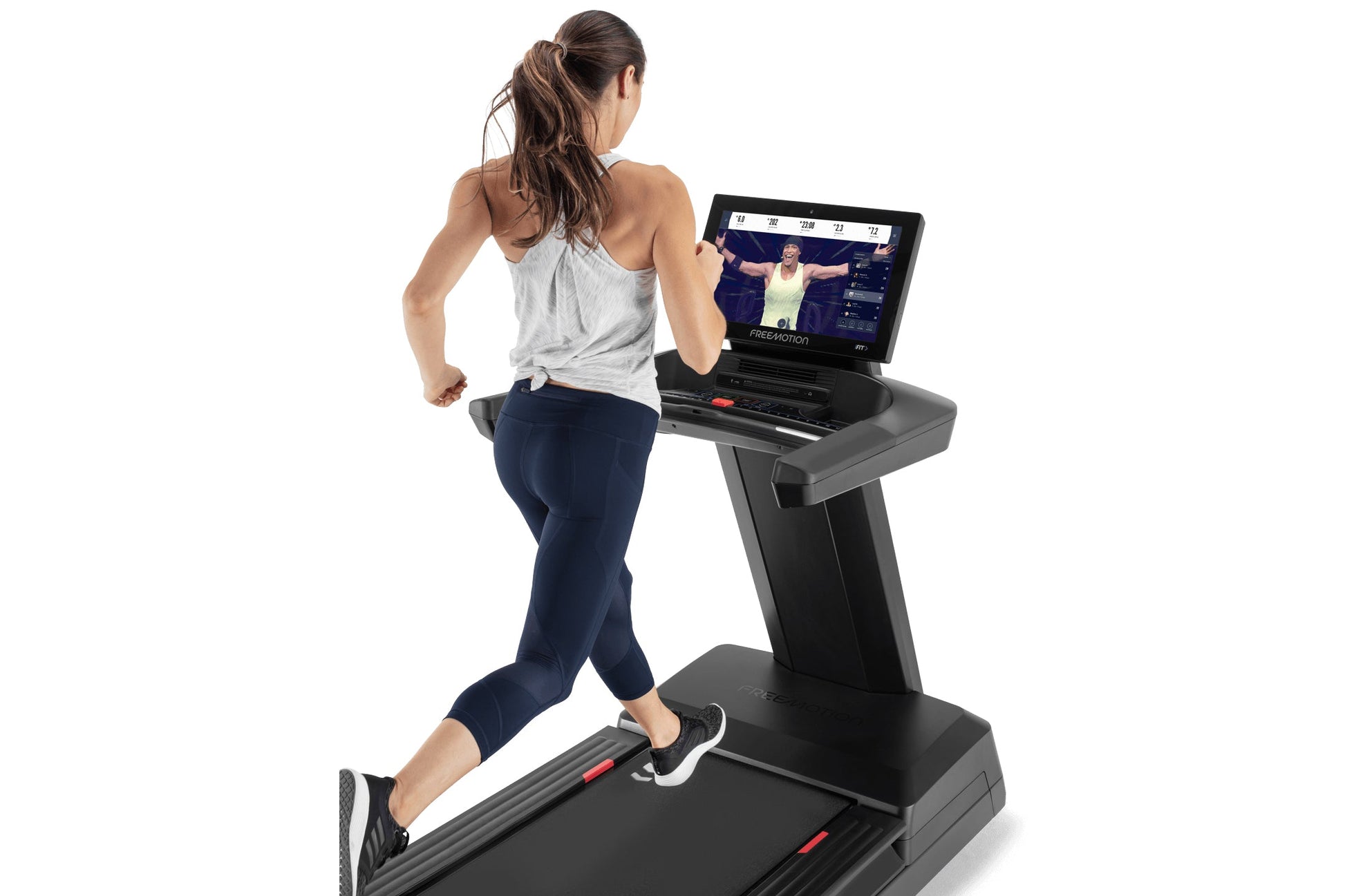 Woman in gray tank top and navy capris running on black Freemotion treadmill with touchscreen display showing workout class i
