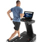 Man running on black Freemotion treadmill with blue touchscreen display showing fitness class, wearing blue t-shirt and black