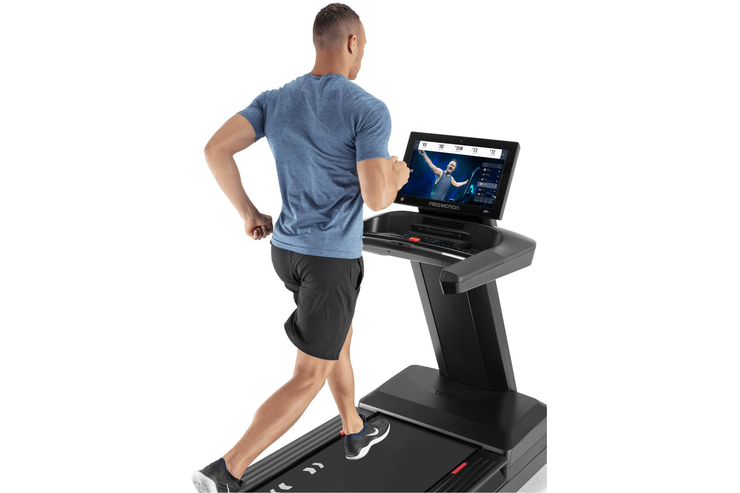Man running on black Freemotion treadmill with blue touchscreen display showing fitness class, wearing blue t-shirt and black