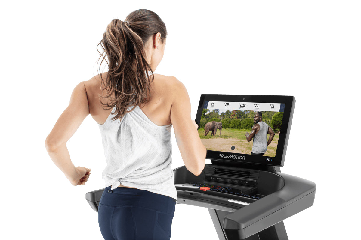 Woman running on Freemotion treadmill with display screen showing workout stats and safari video, worn by model on white back