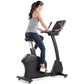 Woman on Freemotion black upright exercise bike with digital display, wearing gray tank top and navy leggings, on white backg