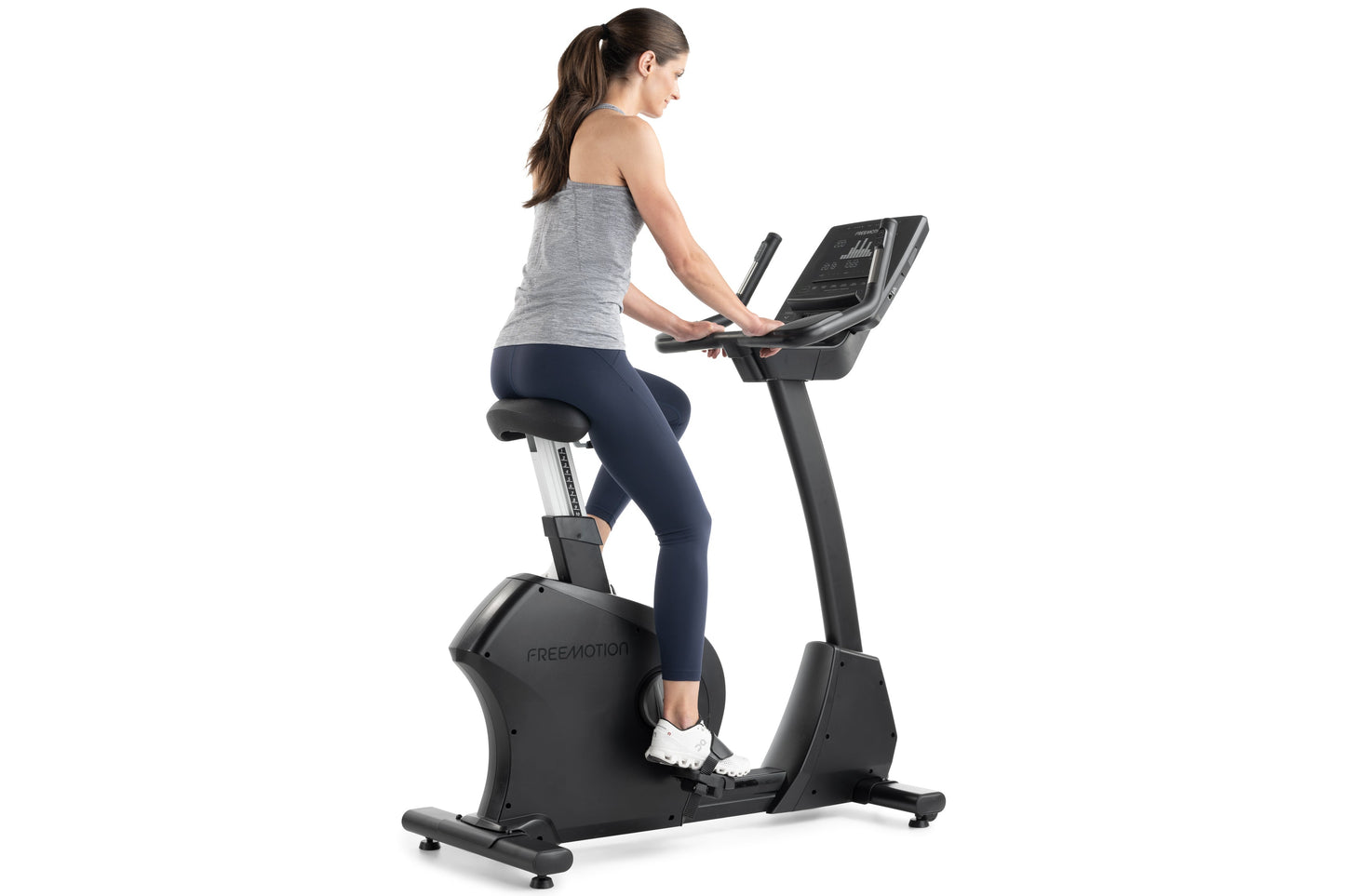 Woman on Freemotion black upright exercise bike with digital display, wearing gray tank top and navy leggings, on white backg