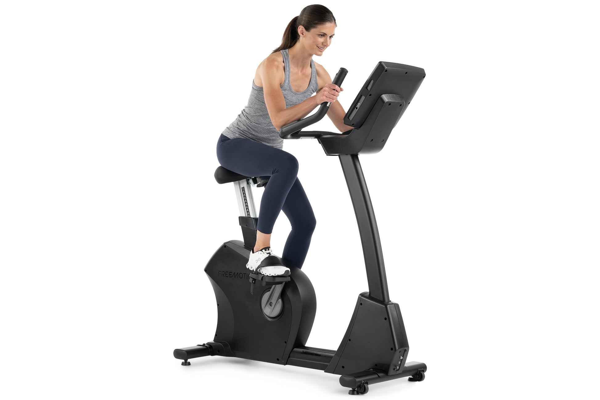 Woman in gray tank top and navy leggings using black Freemotion upright exercise bike with digital display, against white bac