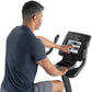 Man using black Freemotion upright exercise bike with touchscreen display showing workout interface, worn by model on white b