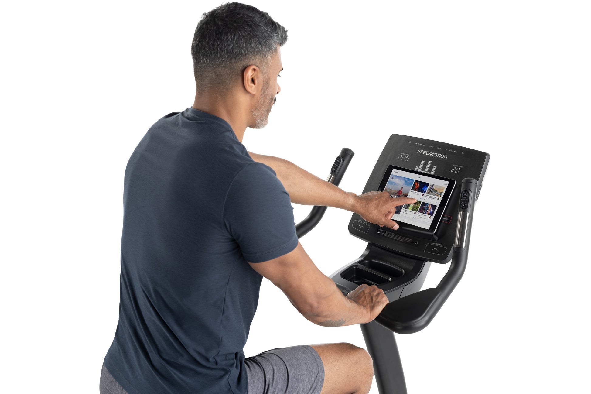 Man using black Freemotion upright exercise bike with touchscreen display showing workout interface, worn by model on white b