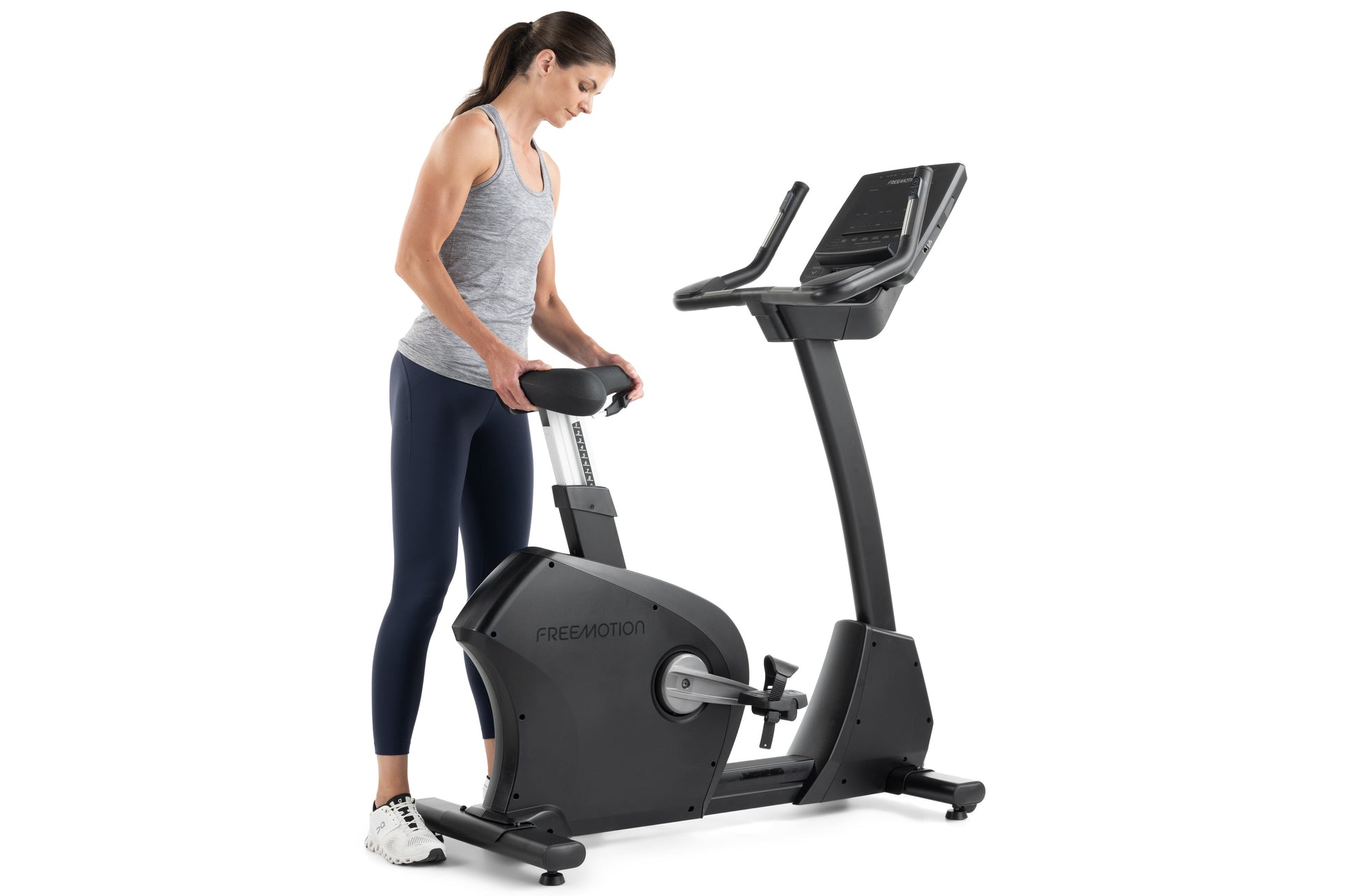 Woman wearing gray tank top and navy leggings using black Freemotion upright exercise bike with digital display on white back
