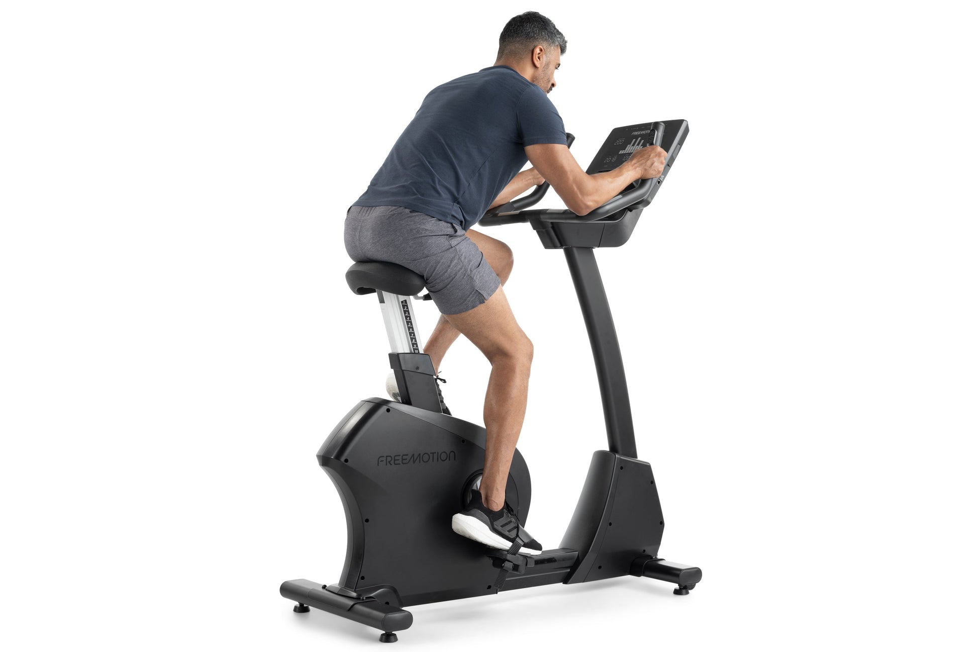 Man using black Freemotion upright exercise bike with digital display, wearing navy t-shirt and gray shorts on white backgrou