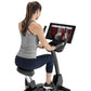 Woman seated on black upright exercise bike with tablet display, wearing gray tank top and navy capris, shown from behind on 