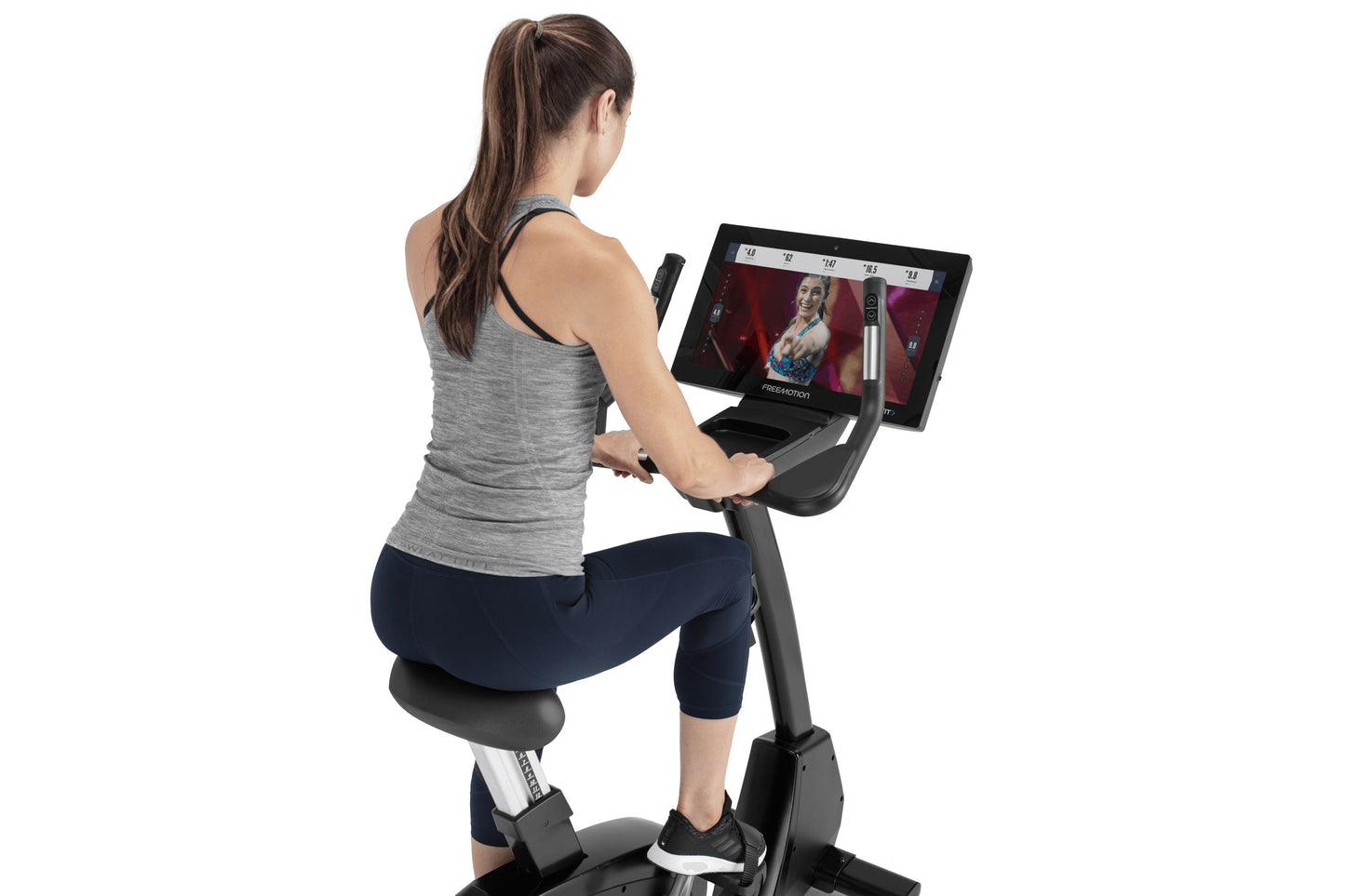 Woman seated on black upright exercise bike with tablet display, wearing gray tank top and navy capris, shown from behind on 