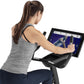 Woman using gray heather racerback tank top on stationary upright exercise bike with touchscreen display showing fitness cont