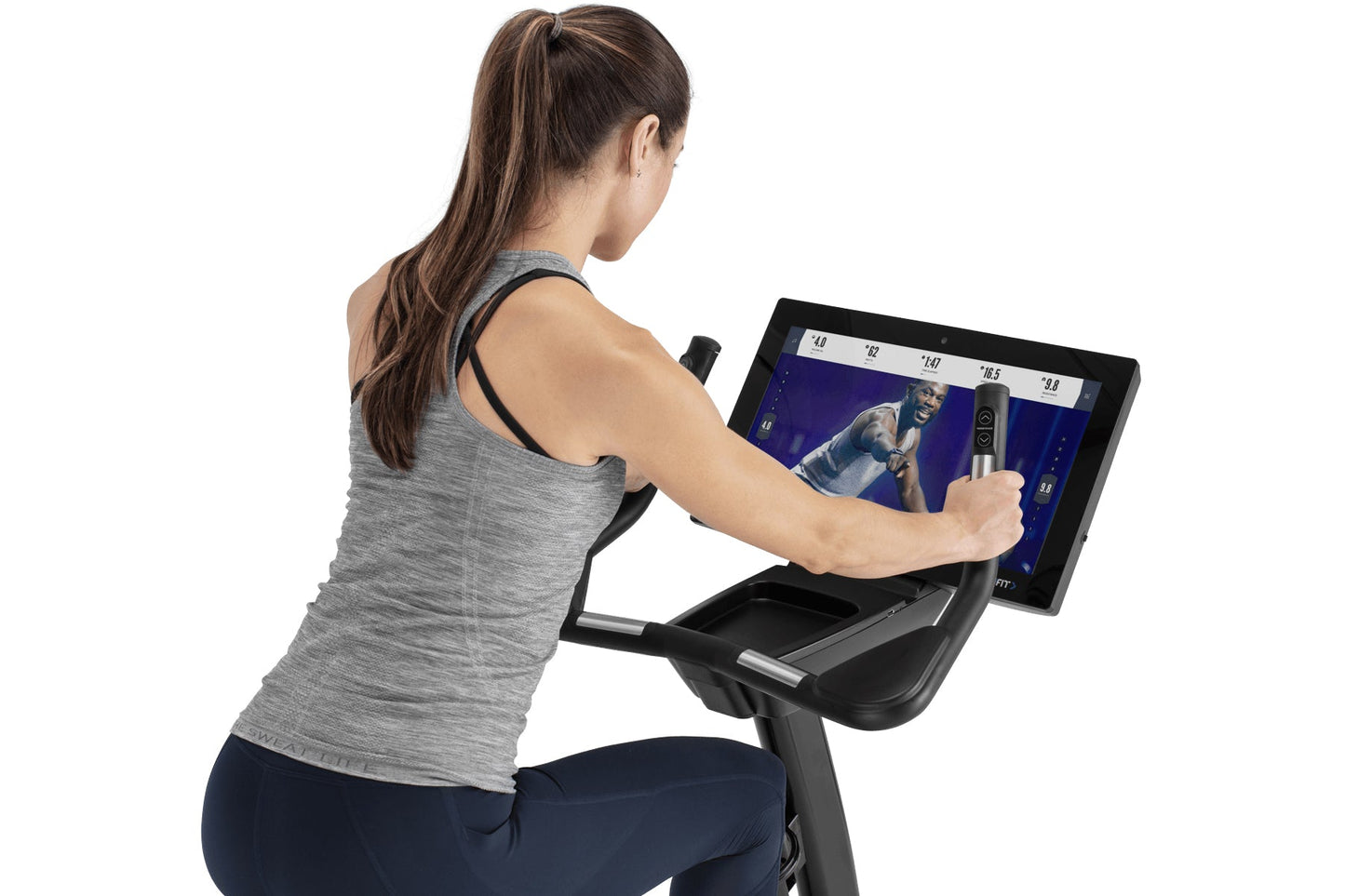 Woman using gray heather racerback tank top on stationary upright exercise bike with touchscreen display showing fitness cont
