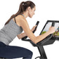 Woman wearing gray sleeveless top and navy leggings using black upright exercise bike with tablet display showing cycling cla