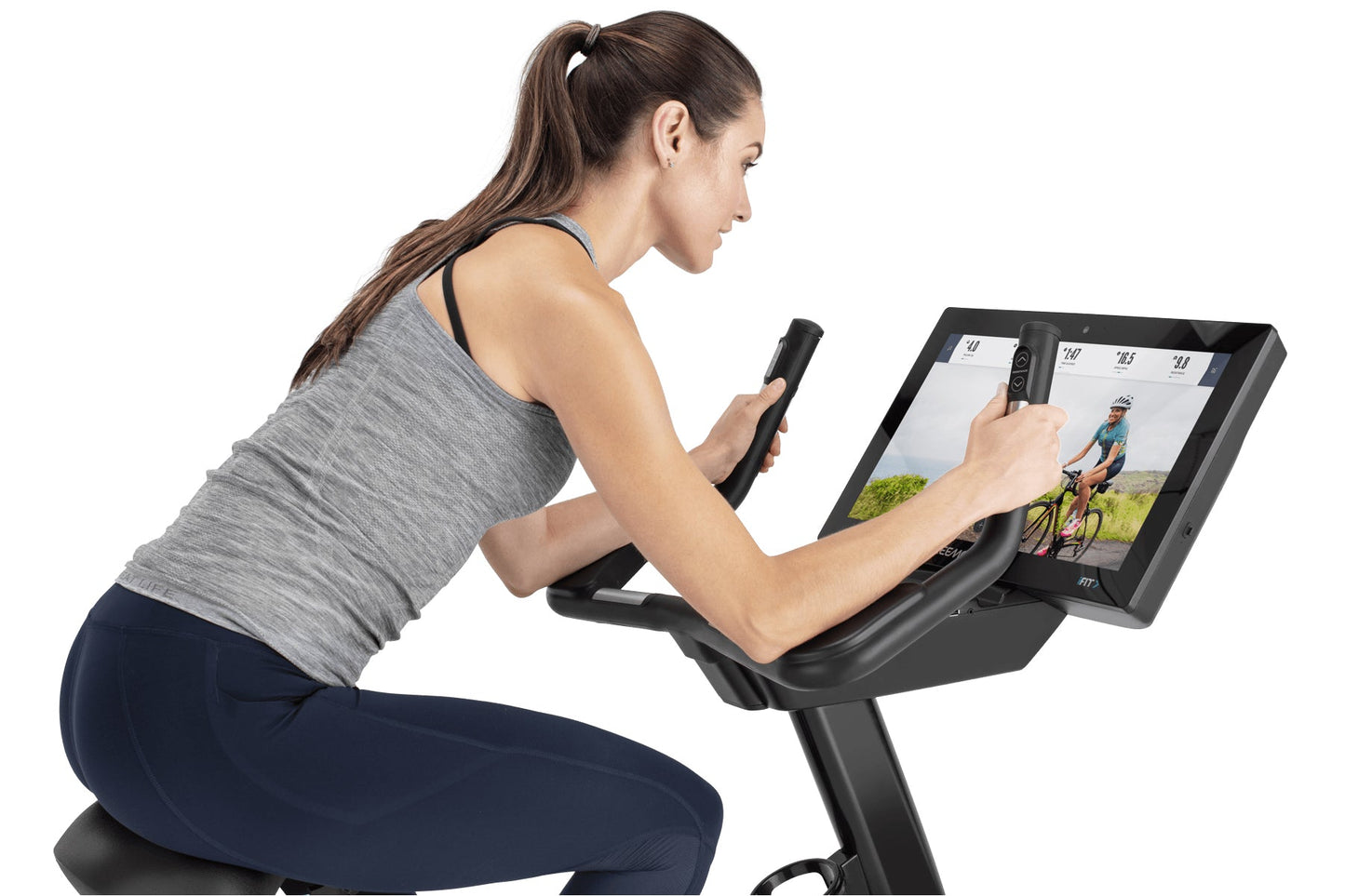 Woman wearing gray sleeveless top and navy leggings using black upright exercise bike with tablet display showing cycling cla