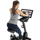 Woman wearing gray tank top and navy leggings using black upright exercise bike with dual tablet screens displaying fitness c