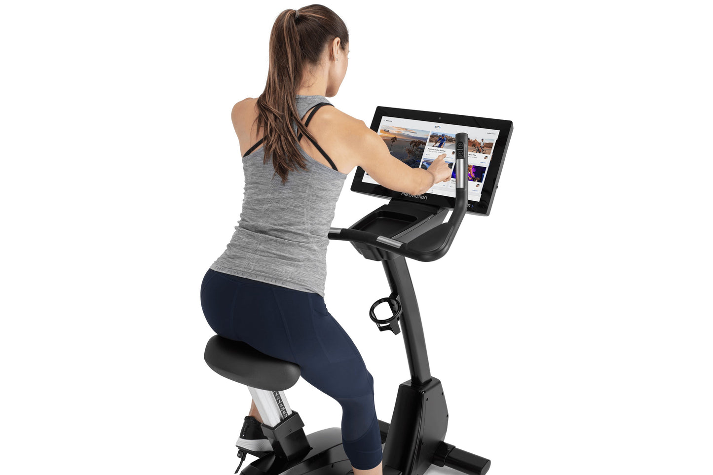 Woman wearing gray tank top and navy leggings using black upright exercise bike with dual tablet screens displaying fitness c