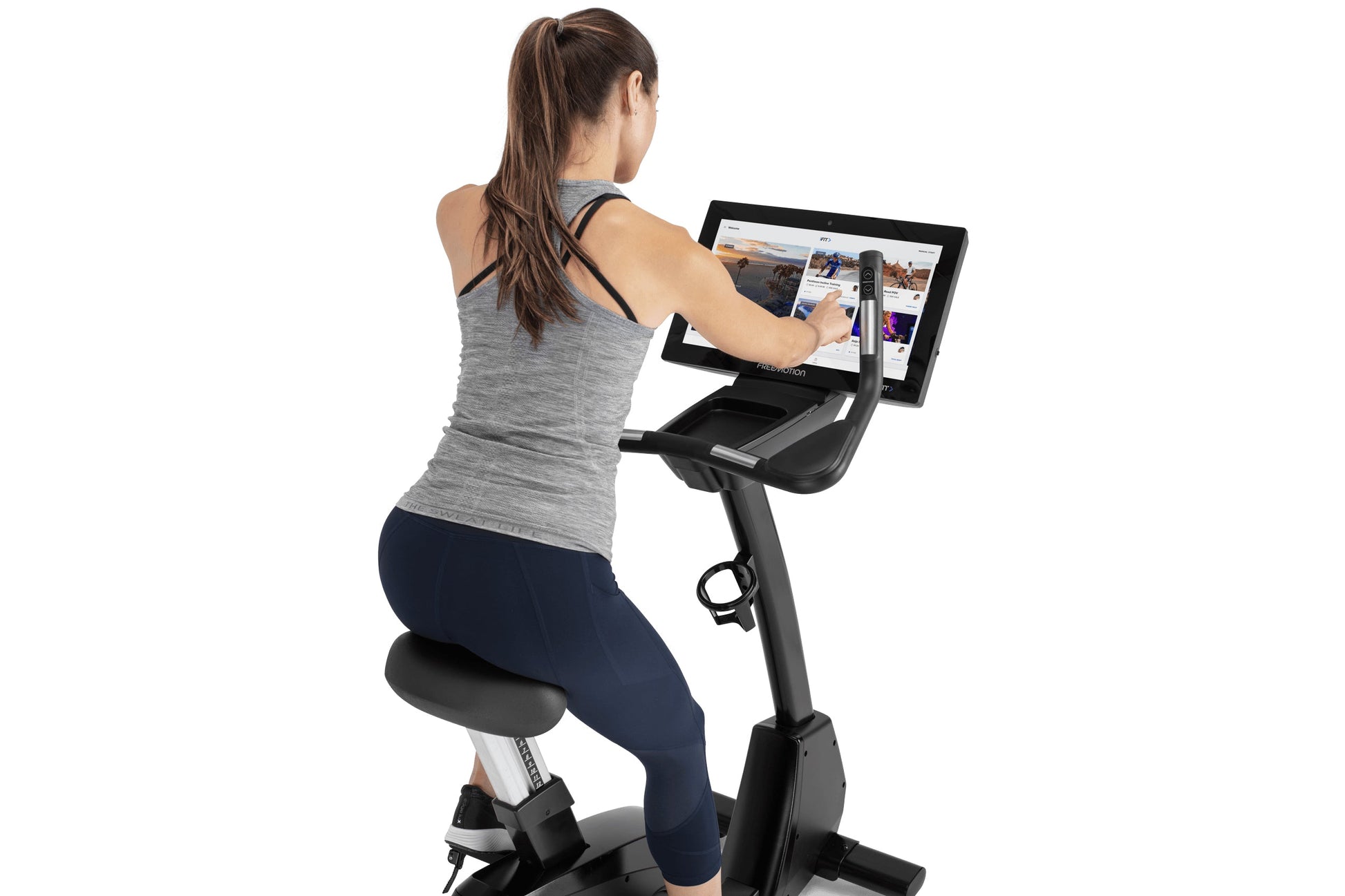 Woman wearing gray tank top and navy leggings using black upright exercise bike with dual tablet screens displaying fitness c