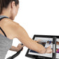 Woman wearing gray sleeveless tank top with black straps using upright exercise bike with digital screen displaying fitness c