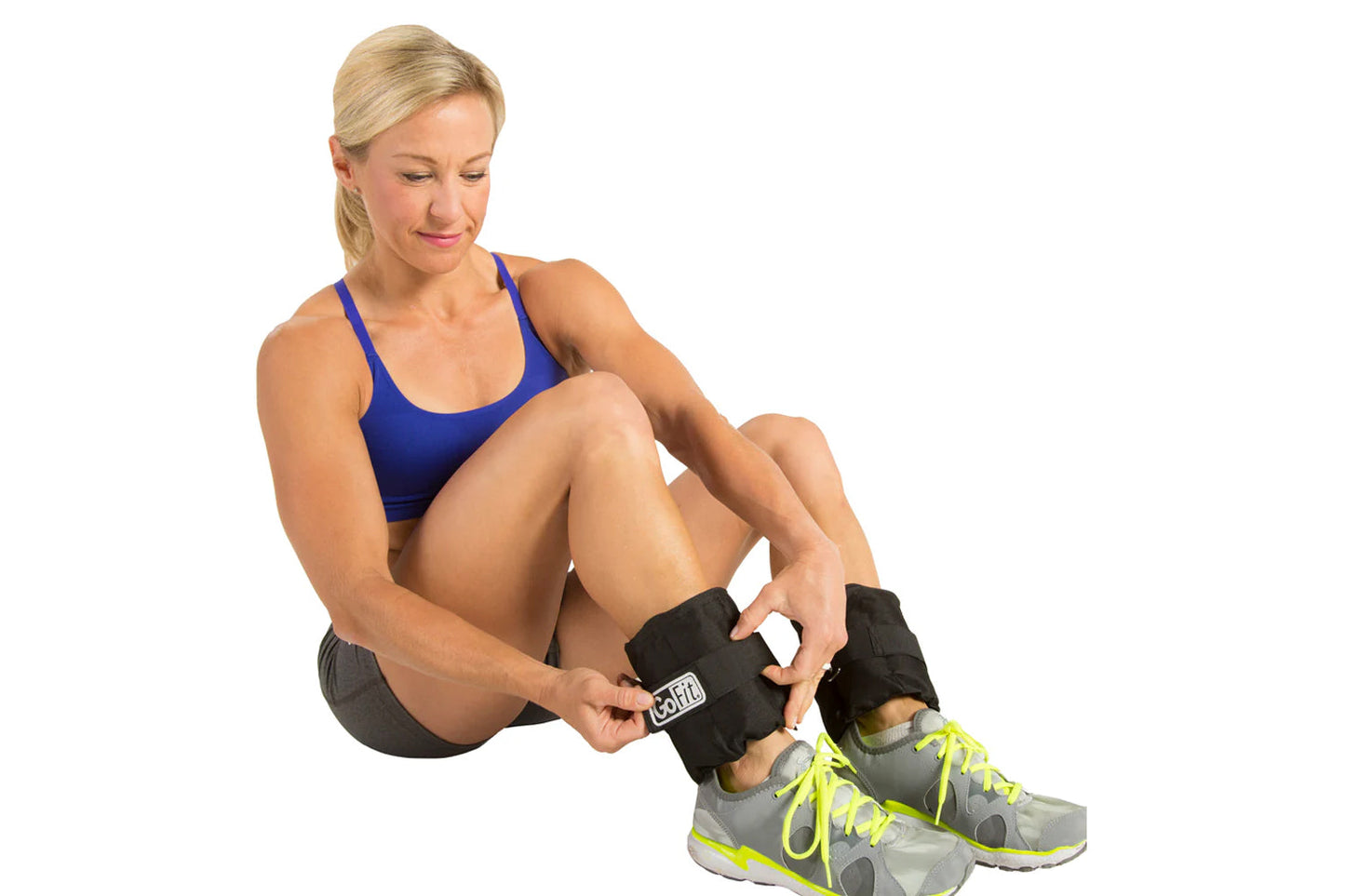 Woman wearing blue sports bra and gray shorts adjusting black GoFit ankle weights with white label, worn with gray and neon y