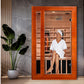 Orange wood far infrared sauna with glass door, woman inside wearing white robe and headwrap, cedar interior visible, potted 