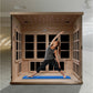 Light wood far infrared sauna cabin with glass doors and interior LED ceiling lights, woman performing yoga stretch on blue m