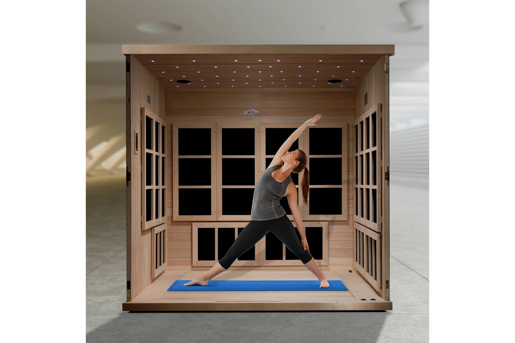 Light wood far infrared sauna cabin with glass doors and interior LED ceiling lights, woman performing yoga stretch on blue m