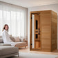 Two-person infrared sauna with golden cedar wood construction and glass door, shown in modern living room with model seated o