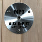 Brushed stainless steel circular control panel with MP3 and AUX Input Max 175mV text, mounted on wood background