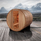 Cylindrical cedar barrel sauna with open door showing wooden interior benches, positioned on wooden deck with snow-capped mou