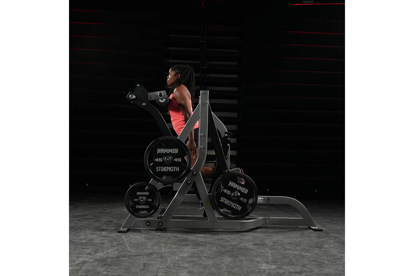 Woman in red tank top using Hammer Strength assisted Nordic hamstring machine with weight plates in dark gym setting
