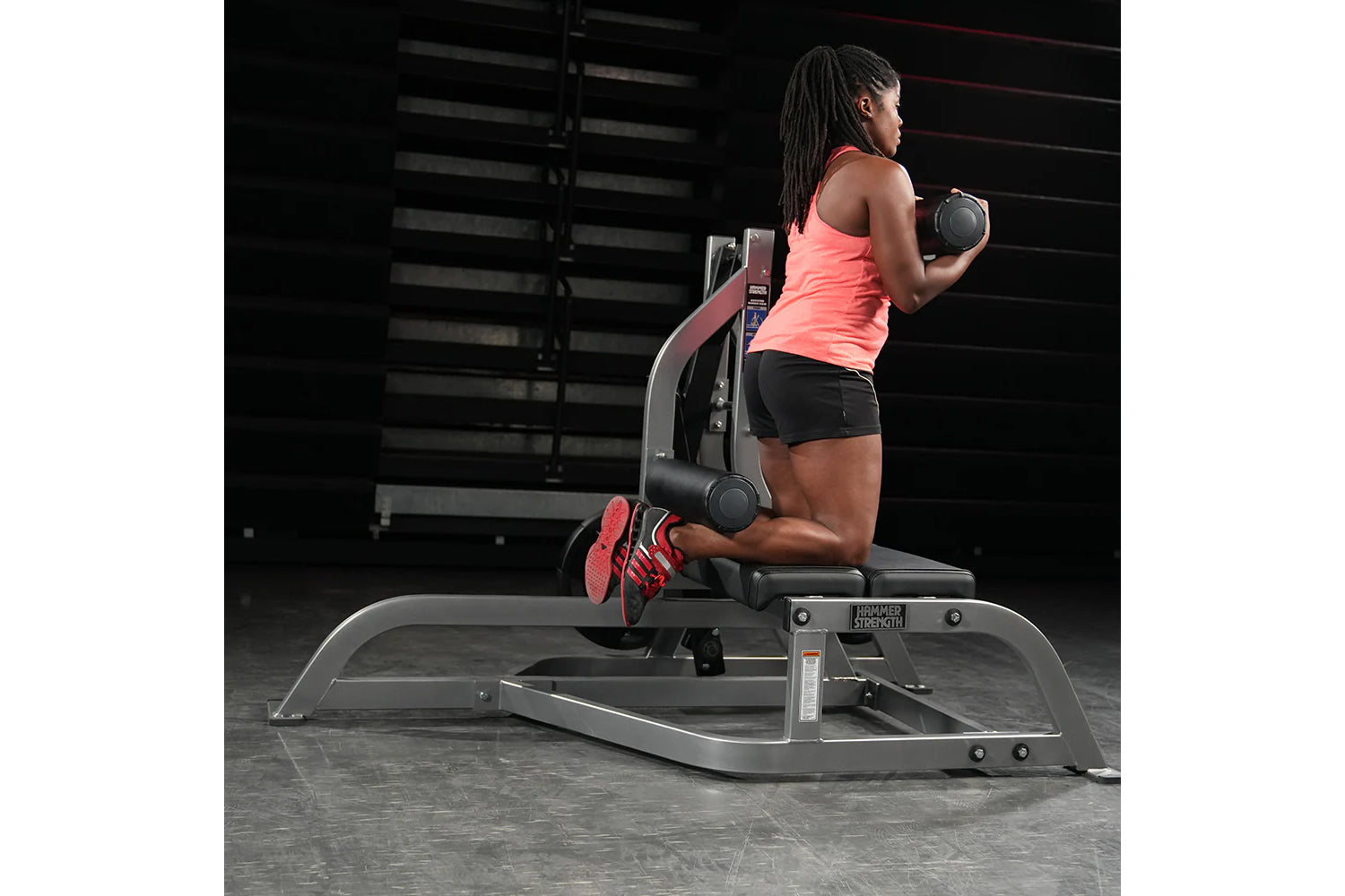 Woman wearing coral tank top and black shorts holding dumbbells on Hammer Strength exercise machine in gym setting