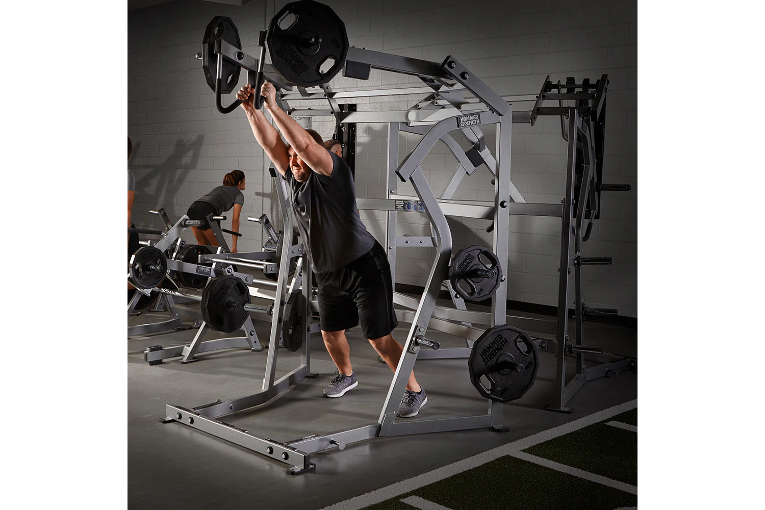 Man performing overhead press with loaded barbell on silver and black Hammer Strength jammer machine in gym setting