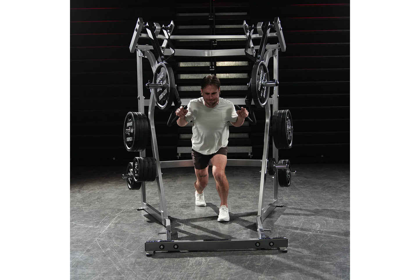 Man wearing light gray t-shirt and black shorts running through metal weight rack machine with loaded barbells in gym setting