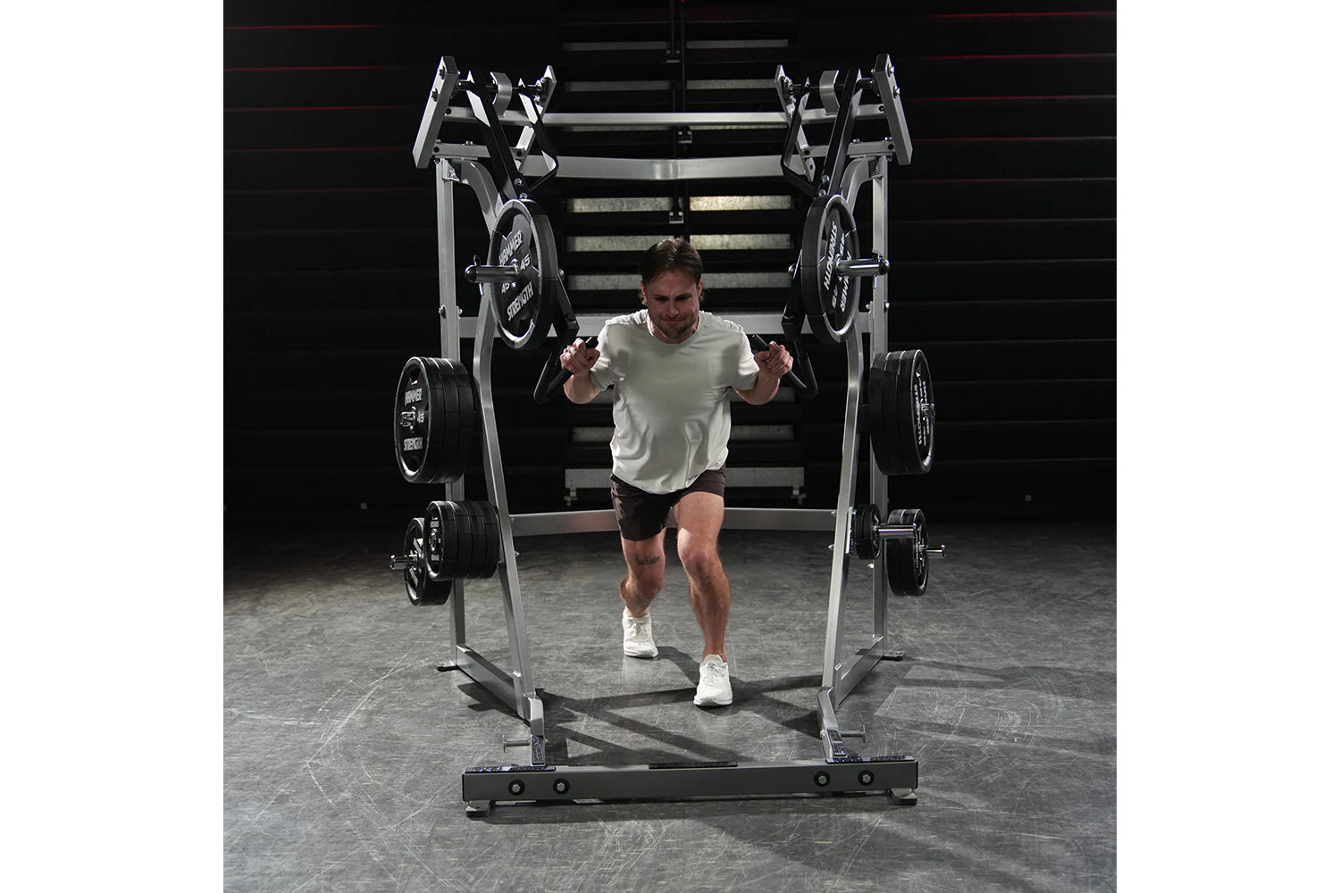 Man wearing light gray t-shirt and black shorts running through metal weight rack machine with loaded barbells in gym setting